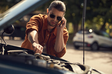 young man in stylish attire calling his insurer and looking at opened engine hood of his car