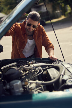 attractive young man in brown shirt with accessories looking attentively at engine of his car