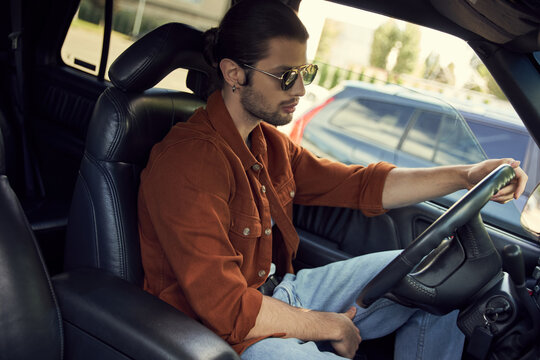 appealing sexy driver with stylish glasses in brown shirt behind steering wheel of his car, fashion