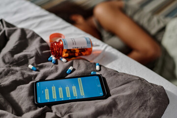 Smartphone with cyberbullying messages on screen and scattered pills on grey blanket covering bed with desperate teenage girl on background