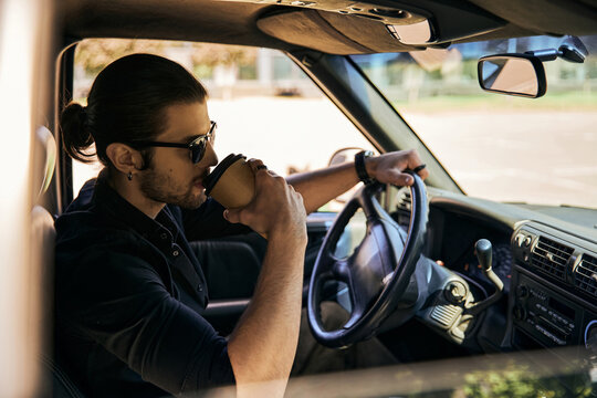good looking stylish male model in black outfit drinking coffee behind steering wheel, sexy driver