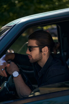 good looking elegant man with earring and beard in black attire behind steering wheel, sexy driver
