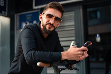 A young happy hipster man with an electric scooter standing in front of a modern business building on the city street and using a mobile phone.