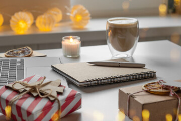 Obraz na płótnie Canvas Notepad with a pen, laptop, coffee cup, wrapped gifts on a desk table. Winter Festive atmospheric mood. Preparation for Christmas. Business Holidays Concept. Freelancer's desktop during Xmas vacation