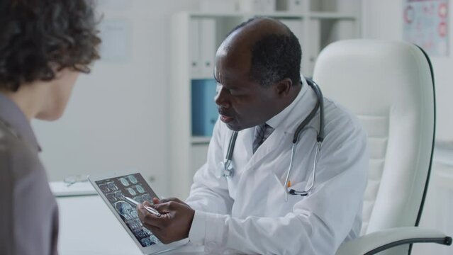Medium side shot of black male physician holding tablet and explaining tomography results to female patient during appointment in health clinic