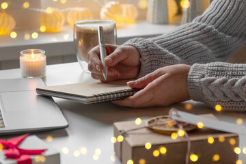 Young woman writing christmas cards, makes order on laptop. Female picking gifts online purchases...
