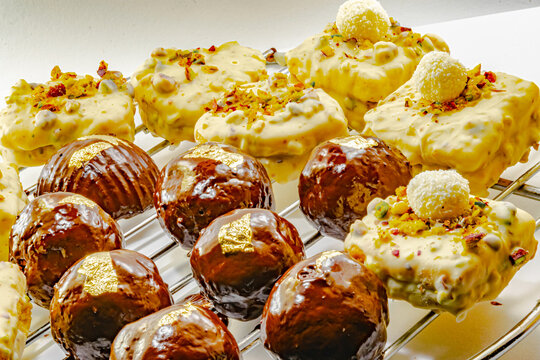 Bniwen, bniouene or bniouen. Non baking Algerian chocolate cakes decorated with a gold leaf and shortbread cakes coated with white chocolate, pistachio chips and coconut powder arranged on a grid.