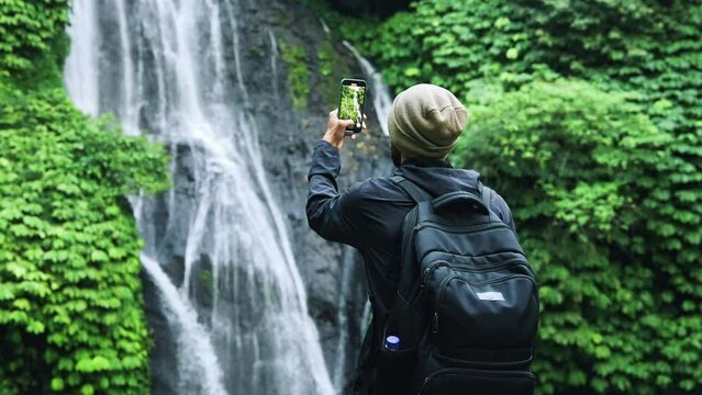 Adult Man Traveling in Green Nature Takes Phone Photo of Forest Waterfall. Lifestyle of Young Unrecognizable Person at Hike Destination of Spring Water Fall. Concept of Traveler Making Journey Videos
