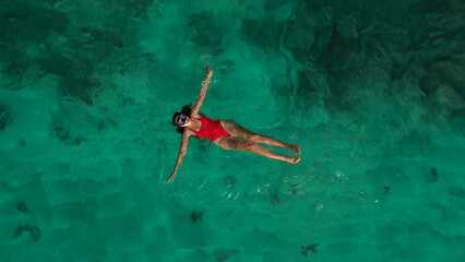 Aerial view of a young woman in a red swimsuit who swims with a mask near the shore in the ocean and watches the coral reef. Snorkeling in the ocean, emerald colored water.