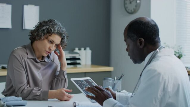 Medium pan shot of Caucasian woman with grey hair complaining about migraine to African American doctor who looking at MRI results on tablet in hospitalMedium pan shot of Caucasian woman with grey hai