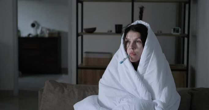 A woman with a cold measures her temperature with a thermometer while wrapping her head in a blanket. Chills and fever from high temperature, treatment, and health care at home.
