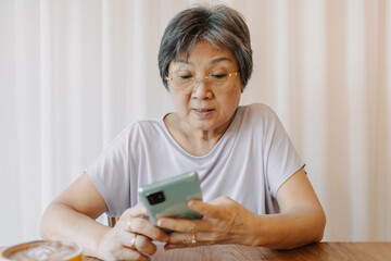 Happy asian old woman smiling and using phone for chatting, reading news and looking at tablet mobile, sitting on chair alone over white curtain.