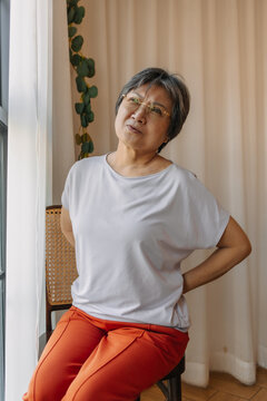 Asian old woman holding and touching back pain, got backache while sitting on chair next to window with painful face.