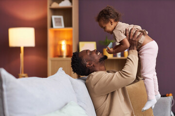 Side view portrait of playful Black father cuddling with little daughter at home and holding her up in air smiling joyfully, copy space