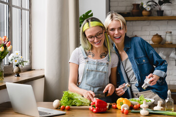Happy young lgbtq female lesbian couple preparing food salad cooking romantic dinner lunch, having fun together at home kitchen while watching culinary recipe show on laptop