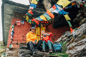 Smiling Backpackers Couple have tea break at small sacred Buddhist monastery decorated multicolored Tibetan prayer flags with mantras. Climbing Mera peak route in Makalu Barun National Park, Nepal
