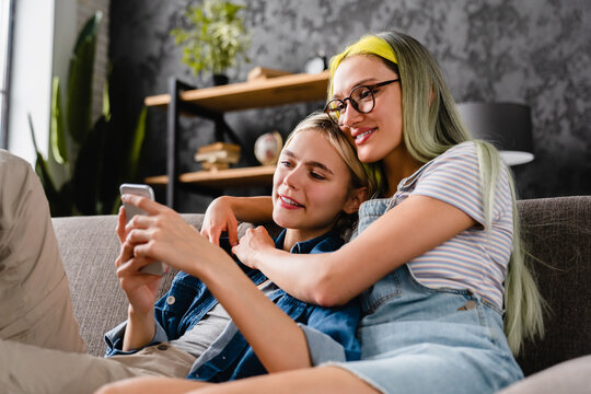 Happy passionate young lesbian lgbtq couple hugging cuddling spending time together, using smart phone for social media, scrolling and posting for followers at home