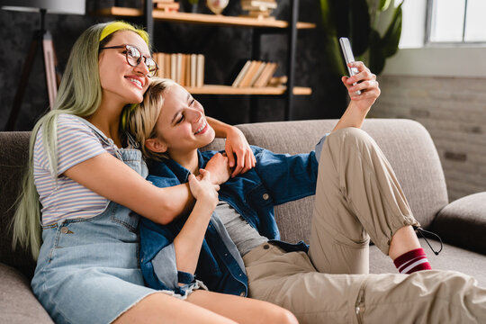 Happy caucasian young female lgbtq lesbian couple hugging cuddling together taking selfie on smart phone for social media, blogging and vlogging at home sitting on the sofa, showing love and care.