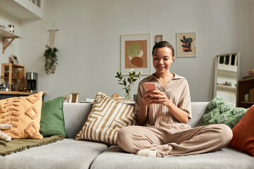 Front view portrait of smiling Black girl using smartphone while sitting cross legged on couch in...