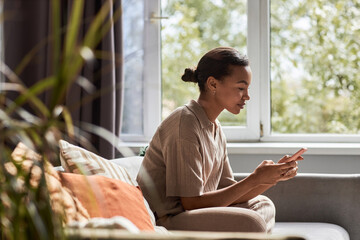 Side view portrait of young Black girl using smartphone while sitting on couch in cozy home, copy...