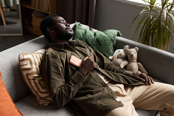 High angle portrait of Black adult man falling asleep on couch at home with smartphone on chest,...