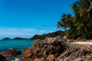Fototapeta na wymiar Blue sea and white sandy beach with tall coconut palm and rocks bright sky.