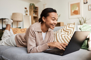 Portrait of smiling Black girl using laptop while lying on couch in cozy home and browsing internet