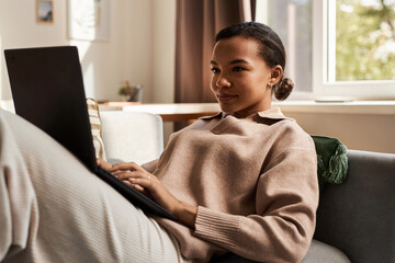 Portrait of elegant Black woman using laptop on couch while relaxing at home during weekend