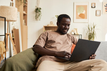 Simple portrait of smiling Black man using laptop at home while relaxing in bean bag, copy space