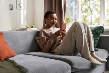 Portrait of young African American woman using smartphone with wireless earphones at home while browsing social media on comfortable couch, copy space