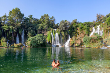 couple swimming in the lake, next to the waterfalls - obrazy, fototapety, plakaty