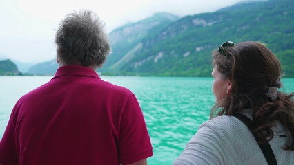 Back of Senior man and adult daughter traveling by boat observing lake and mountain view. Woman pointing at horizon exploring nature with father - obrazy, fototapety, plakaty
