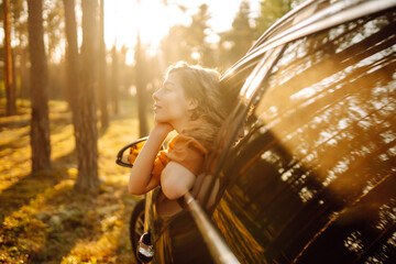 A young woman feels freedom leaning out of a car window in a sunny forest. A traveler enjoys a sunny day from the car window. Travel concept. Active lifestyle.
