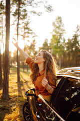 Fototapeta na wymiar A young woman feels freedom leaning out of a car window in a sunny forest. A traveler enjoys a sunny day from the car window. Travel concept. Active lifestyle.