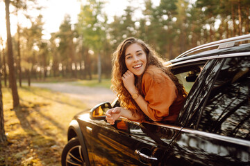 A young woman feels freedom leaning out of a car window in a sunny forest. A traveler enjoys a sunny day from the car window. Travel concept. Active lifestyle. © maxbelchenko