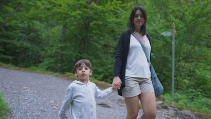 Naklejka na ściany i meble Mother and child holding hands walking in trail path in forest. Mom and son hiking in outdoors, weekend family activity, adventurous journey concept