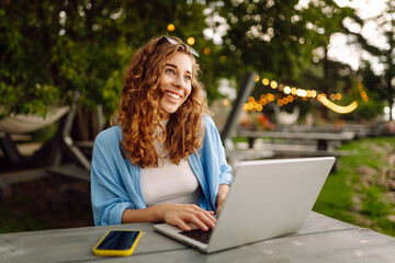 Happy woman in casual clothes working on a laptop in an outdoor cafe near a lake. Young woman freelancer enjoying nature. Tenology concept, working day. - obrazy, fototapety, plakaty