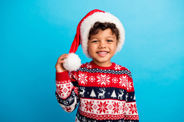 Portrait of toothy beaming little kid with wavy hair wear red pullover santa hat hand touching...