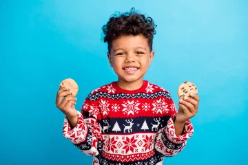 Fotobehang Photo of hungry adorable boy with curly hair dressed ornament sweater hands hold two tasty cookies isolated on blue color background © deagreez