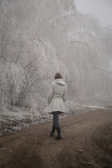 Woman in white coat walking through snowy forest on cold freezing foggy day.