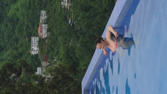 Vertical video. Cheerful young woman enjoying bathing in outdoor pool, amazing view of green hill on the background