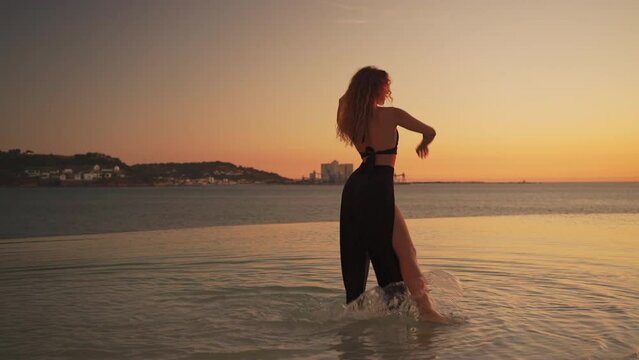 Female dancer moving gracefully at sunset in water. Lady performing dance moves in rays of setting sun. Woman with long wavy hair walking in water and moving rhythmically at sunset