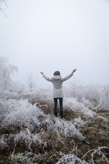 Woman in white coat walking through snowy forest on cold freezing foggy day.