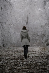 Woman in white coat walking through snowy forest on cold freezing foggy day.