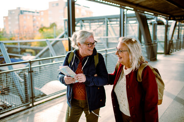 Two elderly women share a laugh while holding a map at a train station