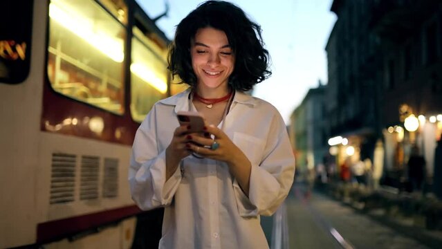 Smiling woman using smartphone on street with night city lights on background. Hipster female blogger typing text message on her cellphone