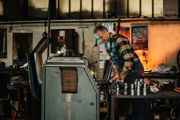 A metal turner leaned over a factory machine, holding knobs. - obrazy, fototapety, plakaty