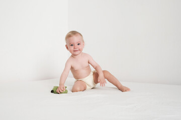 Happy baby in panties is playing with green plush toy on white bed in white room