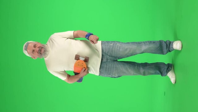 Full body portrait of senior hipster on Chroma key green screen, man in white t-shirt holding skateboard and acts cool. Advertising area, workspace mockup. Vertical video.