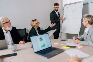 Remote view to confident male manager presents new project plan to colleagues at meeting, explaining ideas on flipchart to coworkers in office. Partners of international company listen to report.